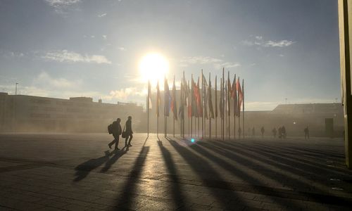Sonne bescheint einen Platz mit Flaggenmasten und zwei Personen, die lange Schatten werfen